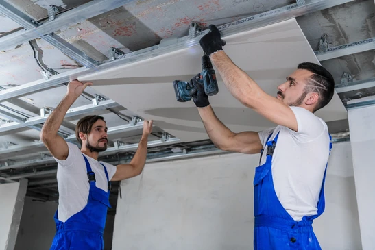 Deux ouvriers en salopettes bleues installent un plafond suspendu dans un local en construction